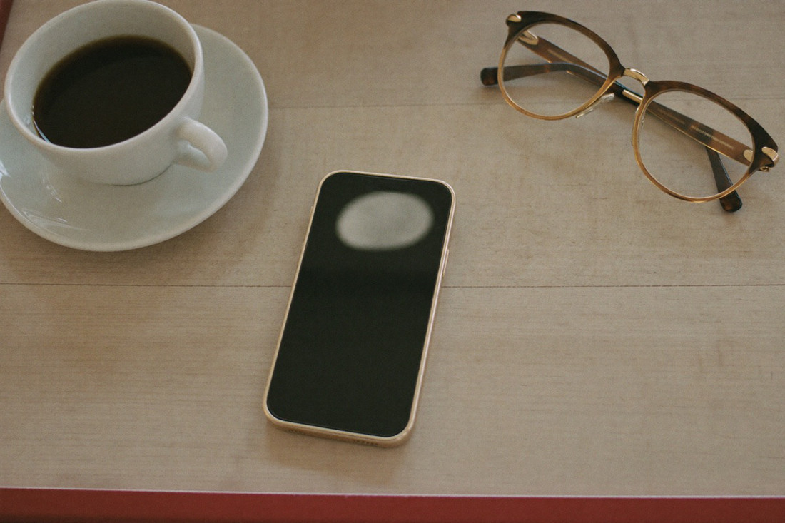 An Android phone with a dark screen on a wooden table beside reading glasses and coffee